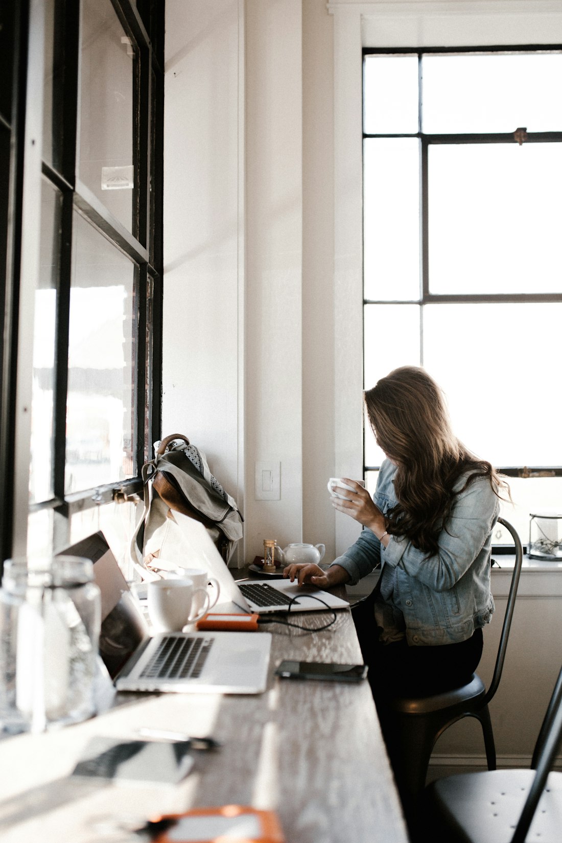 A woman with coffee typing on a laptop by a bright window